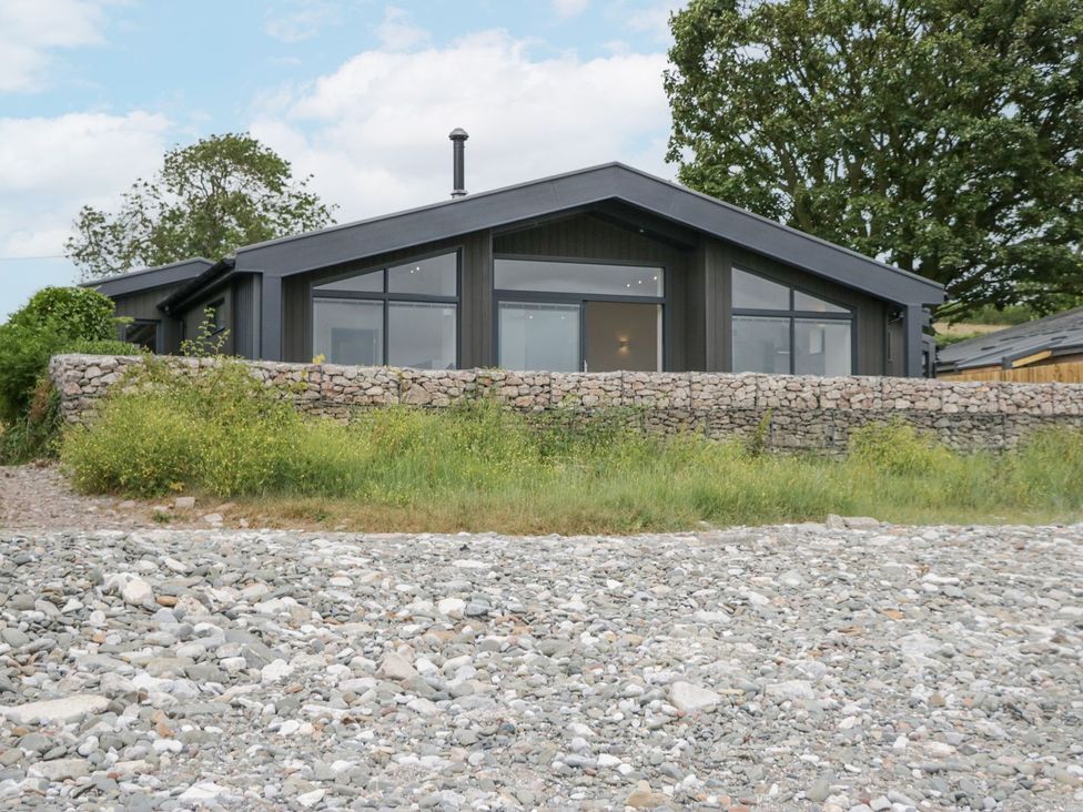 A modern house with large windows behind a stone wall and grass with trees in the background at Driftwood Cottage in Ulverston
