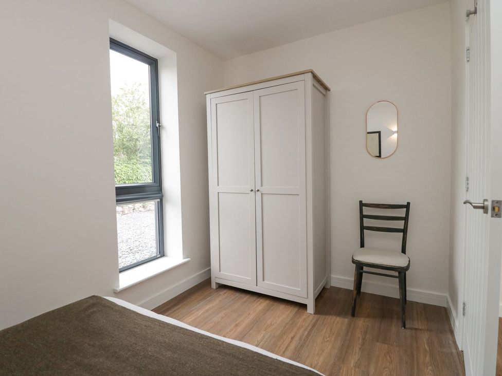 A bedroom corner with a white wardrobe a black chair a small mirror on the wall and a window at Driftwood Cottage in Ulverston