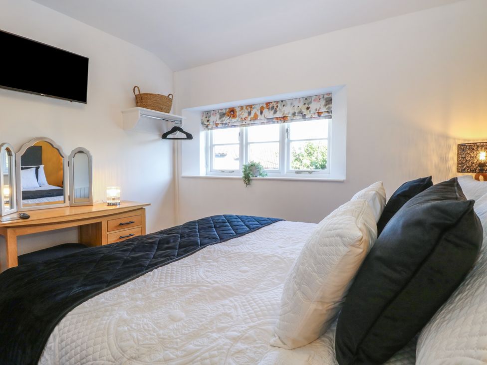 A bedroom with a bed with pillows a wooden dressing table with a mirror a wall-mounted television and a window with a floral blind at Cobble Cottage in Great Ryburgh