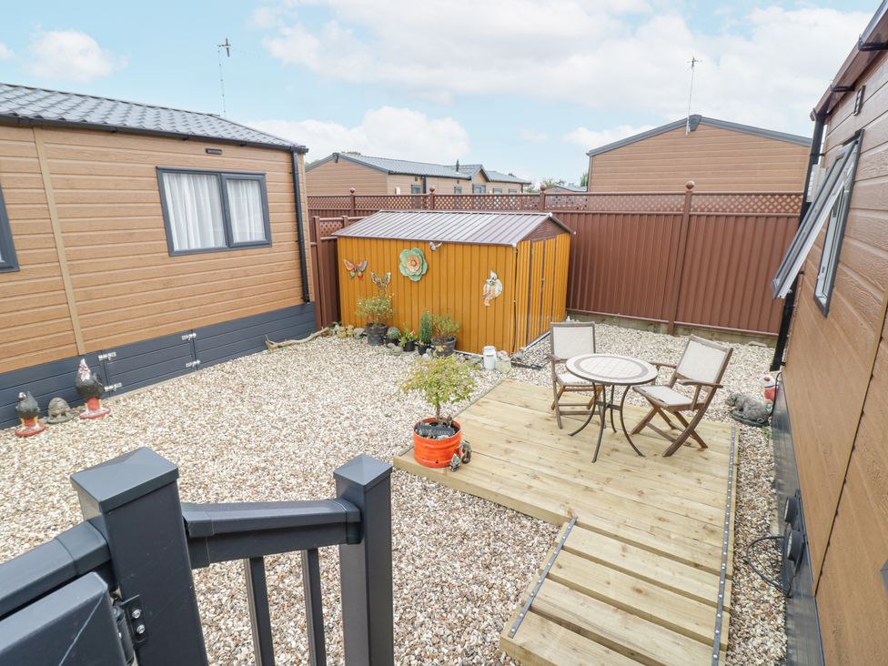 An outdoor patio area with a small wooden deck with a round table and two chairs next to a brown shed and surrounded by gravel at Serenity in Wickhamford near Evesham