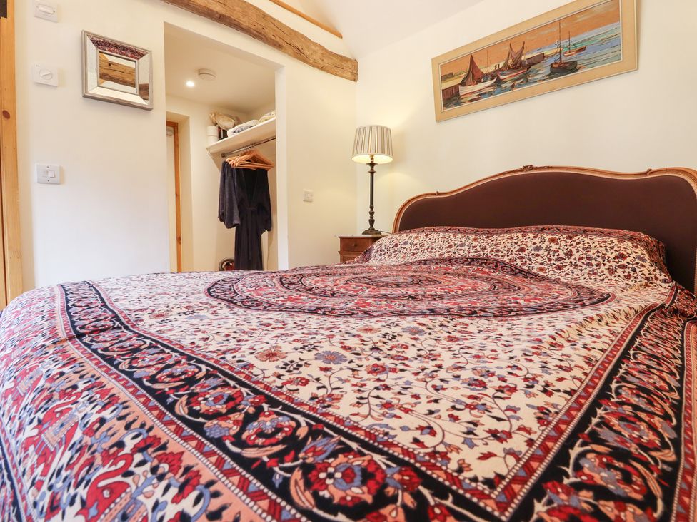 A bedroom with a patterned bedspread a bedside table with a lamp an open closet with clothes and a framed painting above the bed at The Old Stables in Headcorn