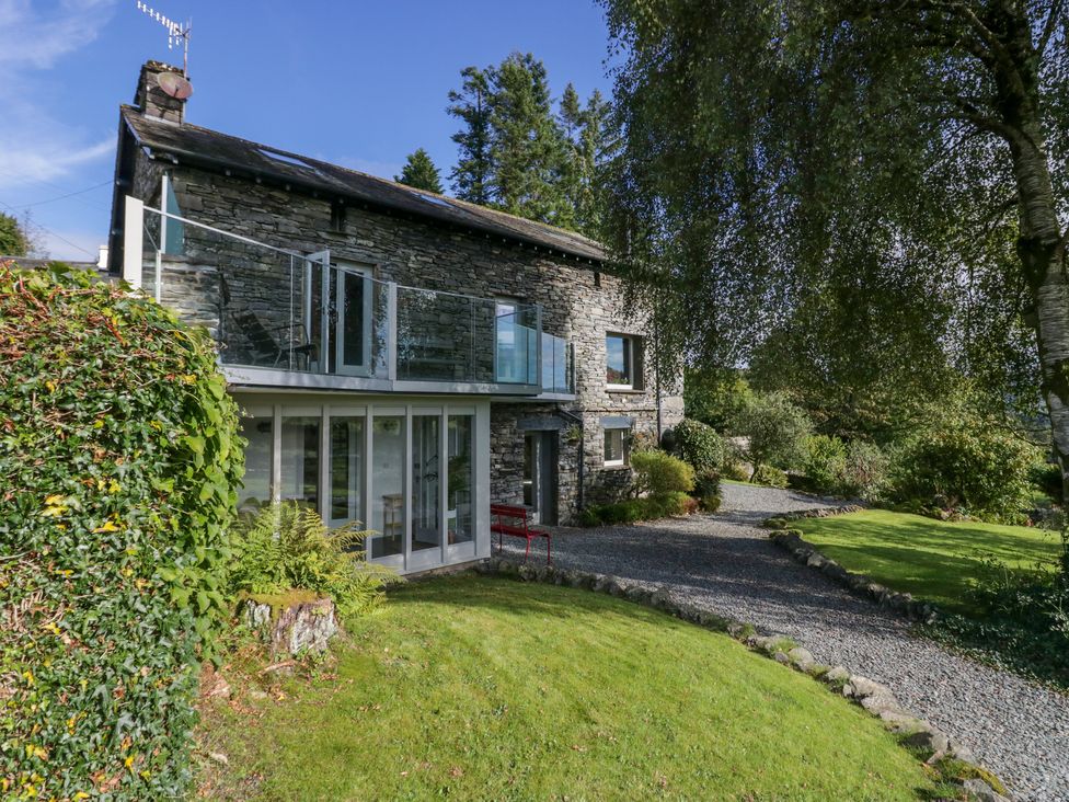 An exterior view of a stone house with glass balcony and gravel path surrounded by trees and lawn at Birkwray Barn Outgate near Hawkshead