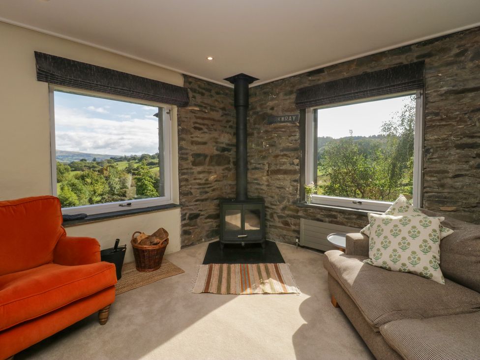 A living room with an orange armchair a brown sofa wood stove and two large windows showing a green landscape at Birkwray Barn Outgate near Hawkshead
