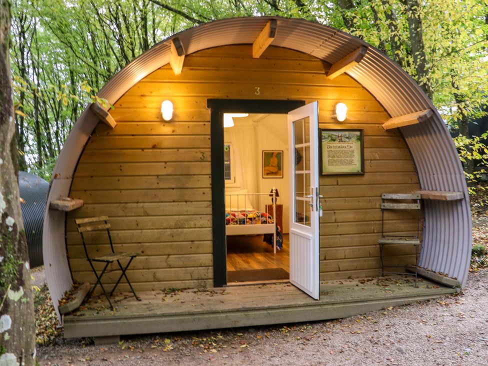 A small wooden curved cabin with an open door showing a bed inside two chairs outside in a wooded area at Ballybeg Pod 7 Ballybeg near Tinahely County Wicklow