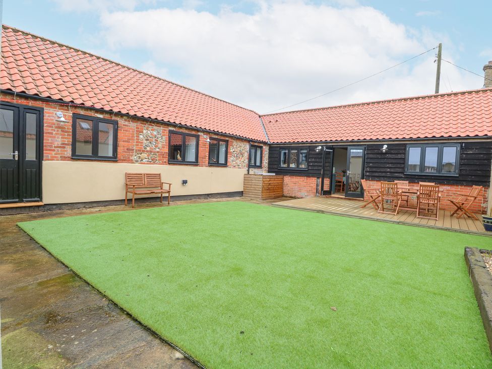 An outdoor patio area with artificial grass a wooden bench a wooden dining table with chairs and brick buildings with tiled roofs