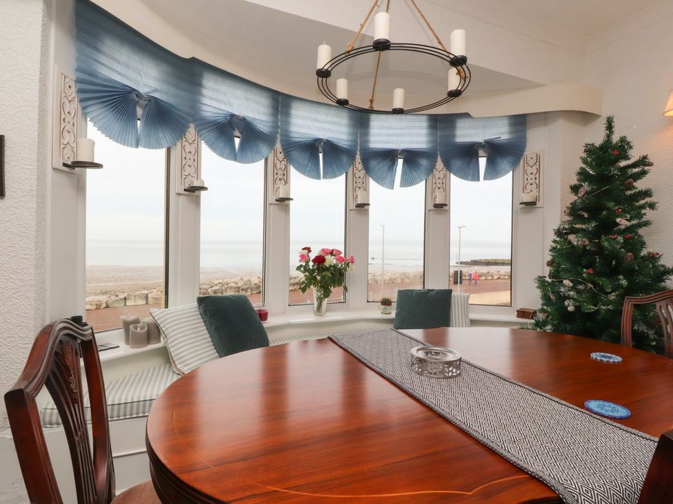 A dining room with a wooden table and chairs a bay window with cushions blue curtains a vase of flowers and a decorated Christmas tree at Love Nest in Heysham