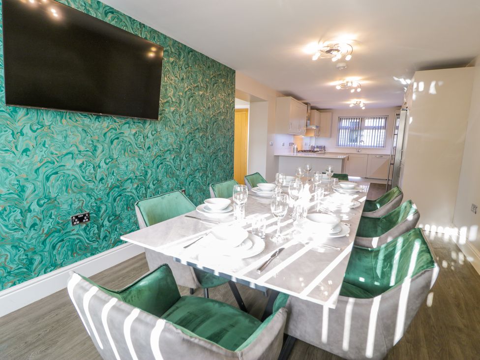 A dining area with a table set with plates and glasses and green and gray chairs beside a kitchen at Bwthyn Y Mor in Tal-Y-Bont