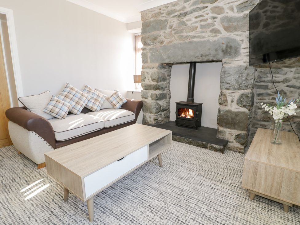 A living room with a sofa with checked cushions a wooden coffee table a stone fireplace with a wood-burning stove and a wooden side table with flowers at Bwthyn Y Mor in Tal-Y-Bont