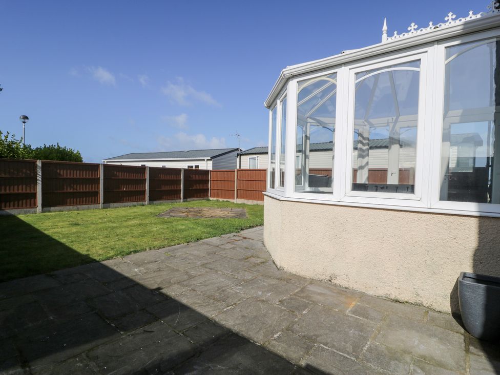 A paved patio area next to a glass conservatory and a grass lawn surrounded by a wooden fence at Bwthyn Y Mor in Tal-Y-Bont