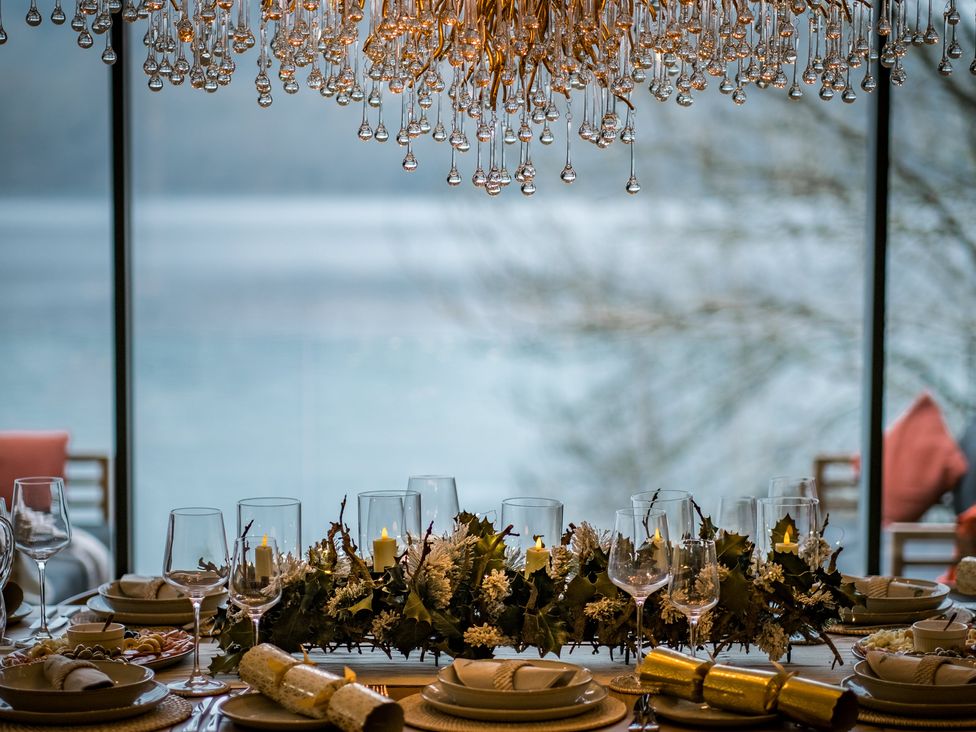 A dining table set with plates glasses candles and festive decorations by a window overlooking water at The Ridge in Bowness-On-Windermere