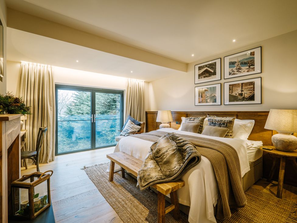 A bedroom with a large bed wooden bench bedside tables lamps and glass doors showing trees outside at The Ridge in Bowness-On-Windermere