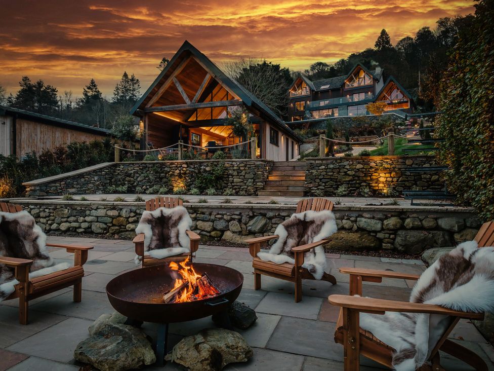 An outdoor patio with wooden chairs and a fire pit in front of stone terrace and cabins at The Ridge in Bowness-On-Windermere