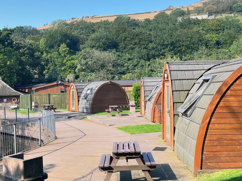 A row of wooden glamping pods with picnic tables outside near a wooded area at Dovestone Pod 20 in Greenfield