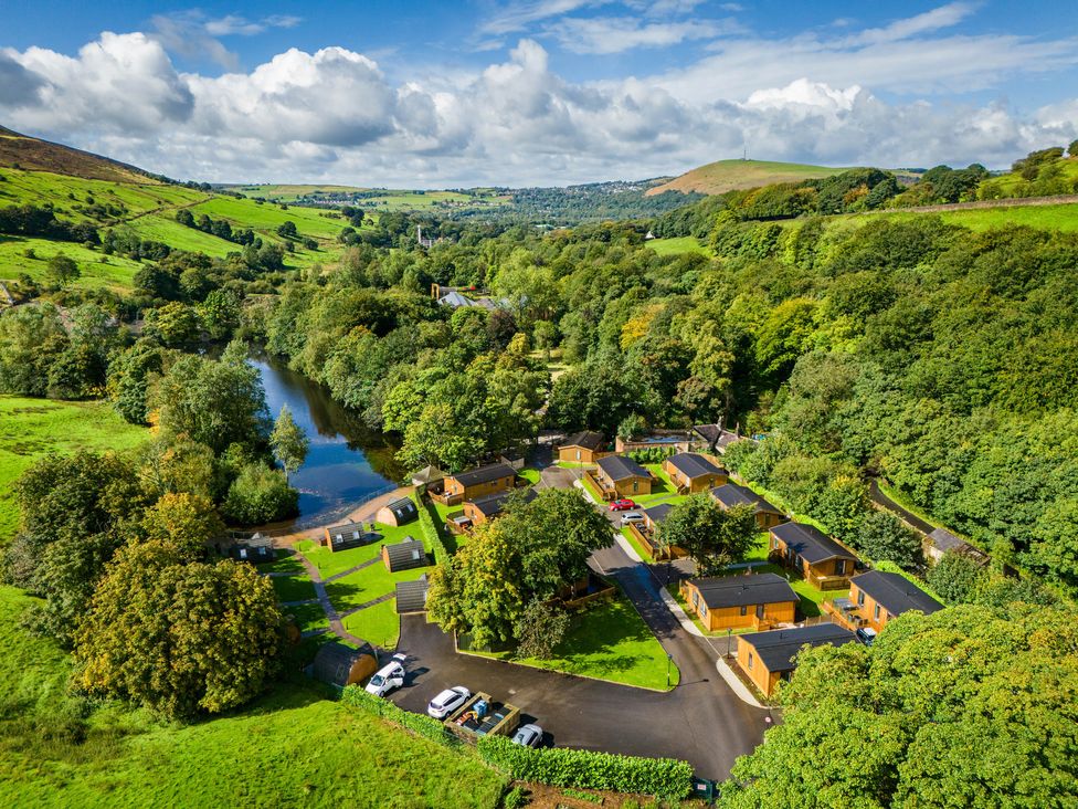 An aerial view of wooden cabins and small pods near a river surrounded by green hills and trees at Dovestone Pod 22 in Greenfield