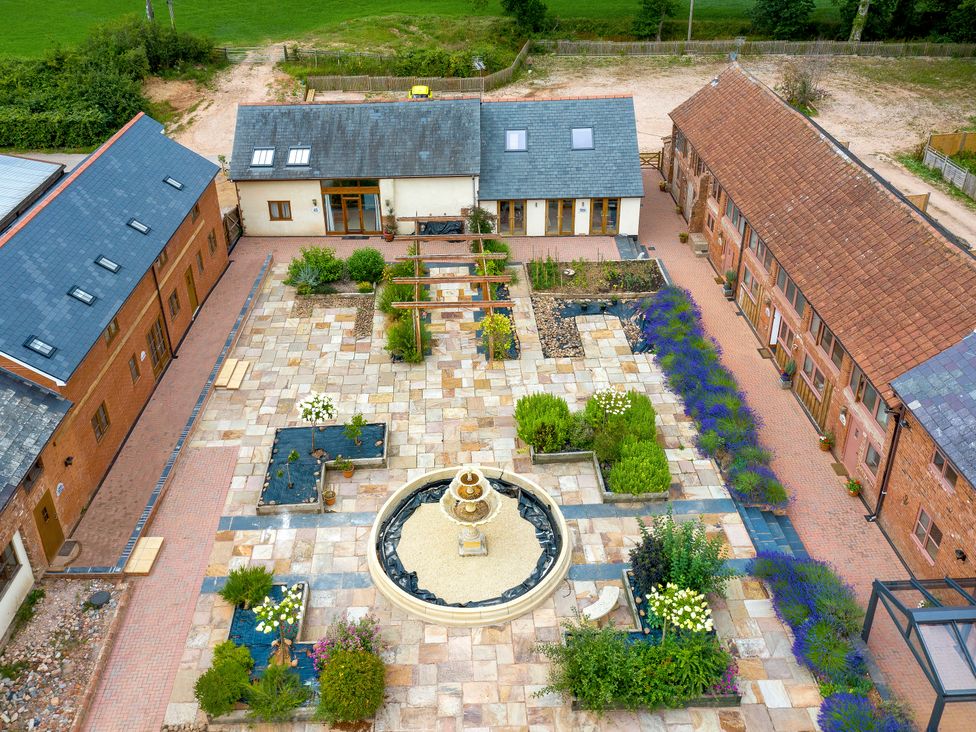 A courtyard with a circular fountain in the center surrounded by flower beds and pathways between brick buildings at Poppy Cottage in Aylesbeare