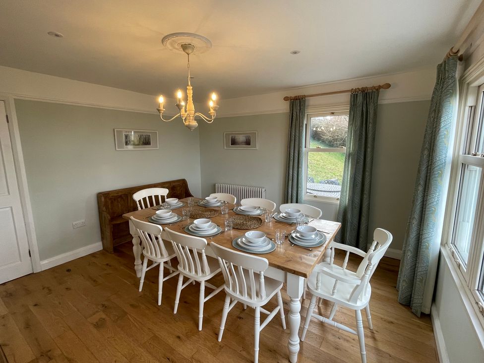 A dining room with a wooden table set for eight people with white chairs and windows with curtains at Bryn Awelon in Harlech