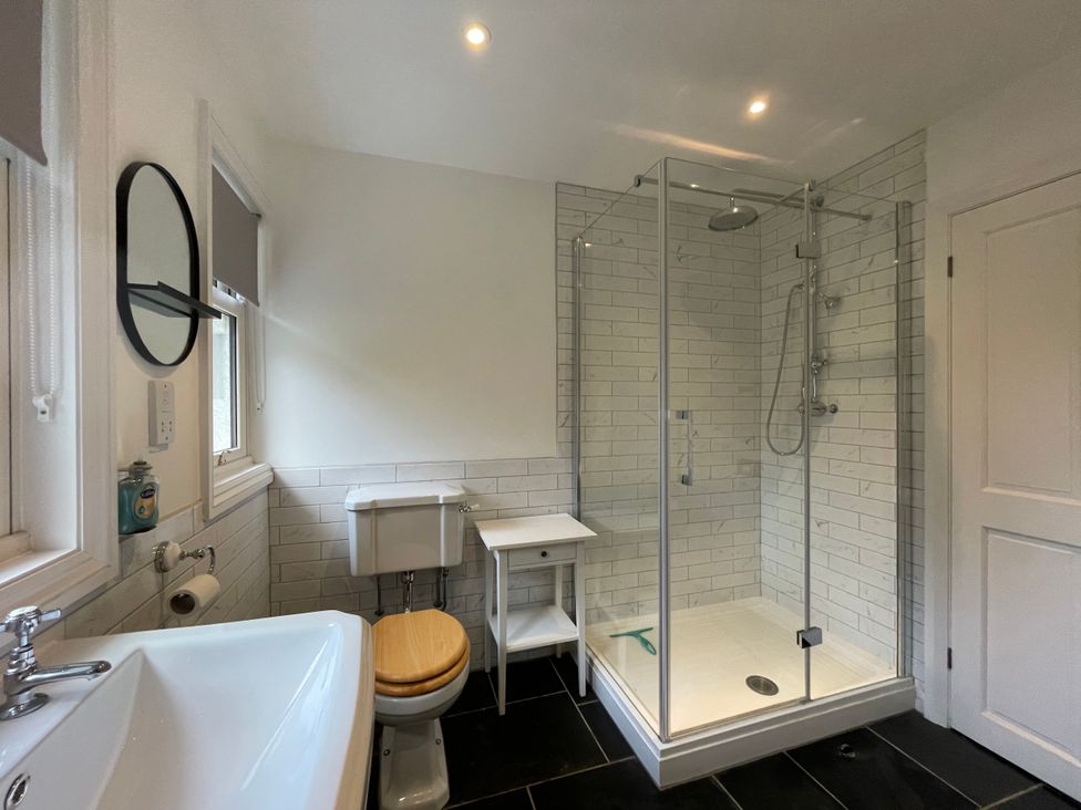 A bathroom with a sink, toilet with wooden seat, glass-enclosed shower, small white table, and a round mirror at Bryn Awelon in Harlech