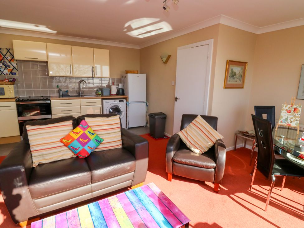 A living area with a brown leather sofa and armchair with striped cushions a colorful coffee table a kitchen with cream cabinets and appliances and a dining table with black chairs at Rosa in Torquay
