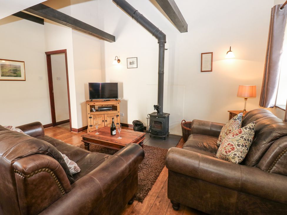 A living room with leather sofas a wood burning stove a wooden coffee table with wine glasses and a bottle a television on a wooden cabinet and framed pictures on the walls at Pencroft Cottage in Carperby