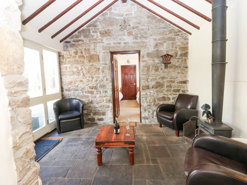 A sitting area with three leather chairs a wooden coffee table a bottle and glasses stone walls and a wood stove at Pencroft Cottage in Carperby