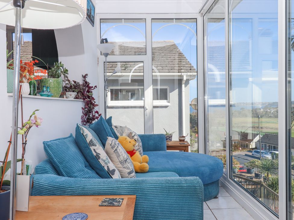 A sunroom with a blue chaise sofa with cushions and a teddy bear near glass doors and windows overlooking a neighborhood at House of Emma in Paignton
