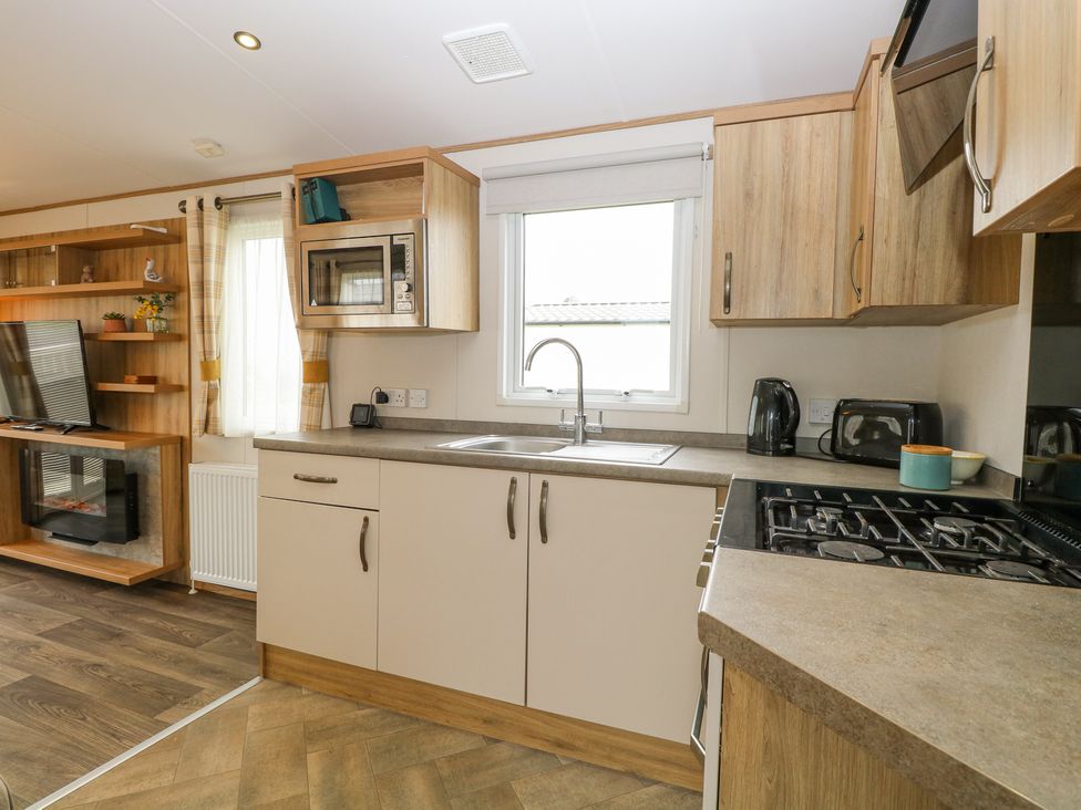 A kitchen with wooden cabinets a microwave a sink a stove a kettle a toaster and a living area with a television and shelves at 75 Foxburrow Hang in Fritton