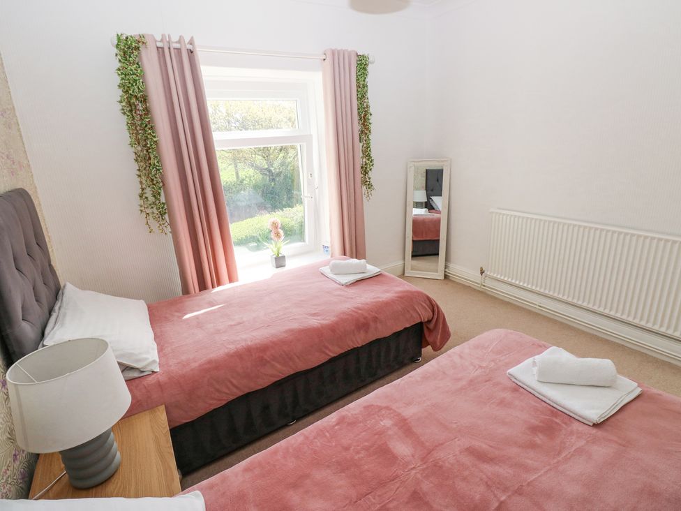A bedroom with two single beds with pink covers a window with pink curtains a wooden side table with a lamp a standing mirror and a radiator at Burry House in Llanmorlais