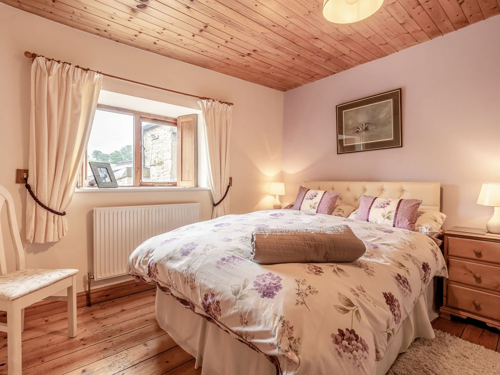A bedroom with a bed covered with floral bedding two bedside tables with lamps a wooden chair a window with curtains and wooden ceiling at Brant View in Cautley near Sedbergh