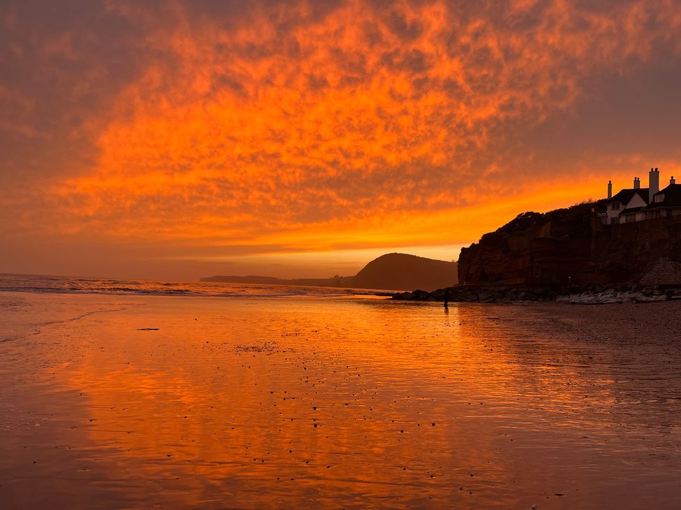 A beach with wet sand reflecting a bright orange sunset sky and houses on a cliffside