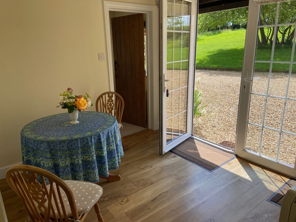A dining area with a round table covered with a blue tablecloth and wooden chairs near glass doors opening to a garden at Oke Apple Cottage in Okeford Fitzpaine