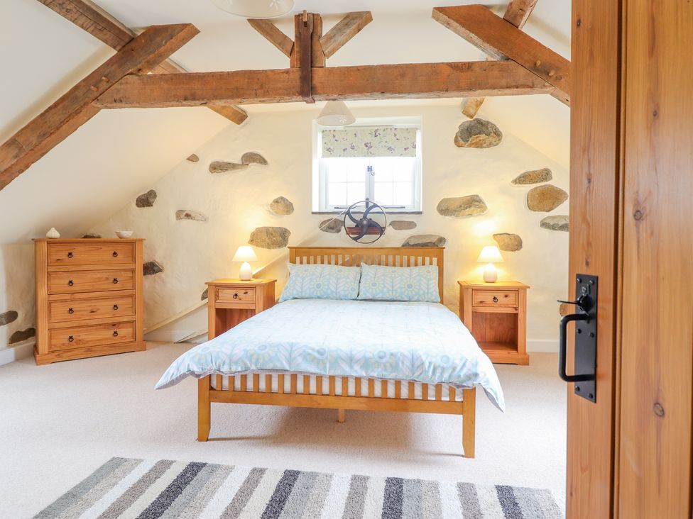 A bedroom with a wooden bed and two bedside tables with lamps a wooden chest of drawers exposed wooden beams and stone accent walls at Ysgybor Yd in Chwilog near Criccieth