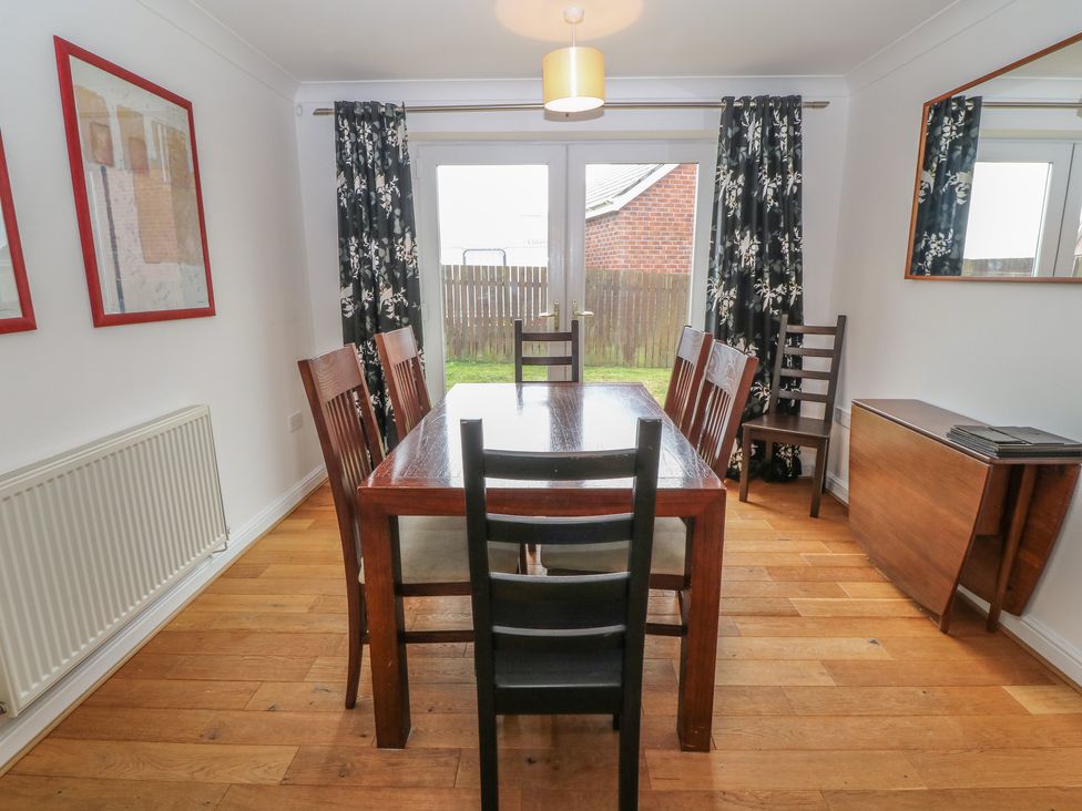 A dining room with a wooden table and six chairs near glass doors with curtains at 5 Cae Derwydd in Cemaes Bay