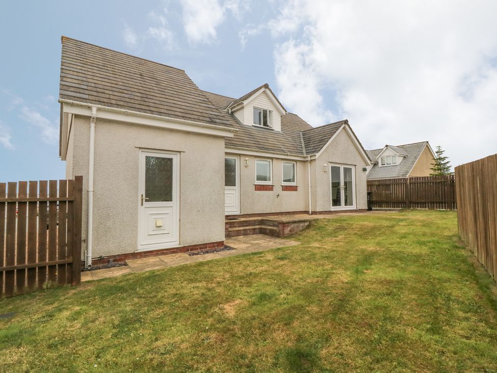 A backyard with grass and a small patio area behind a detached house with a tiled roof and white doors at 5 Cae Derwydd in Cemaes Bay