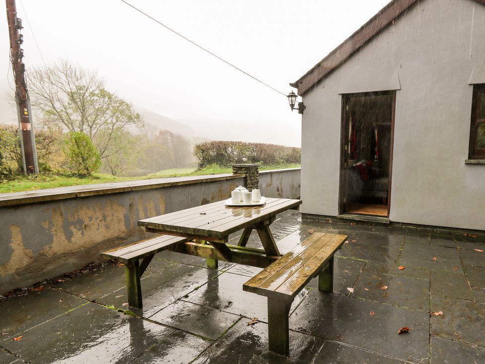 A wet patio with a wooden picnic table and benches next to a house with an open door at Penlon in Capel Bangor near Aberystwyth