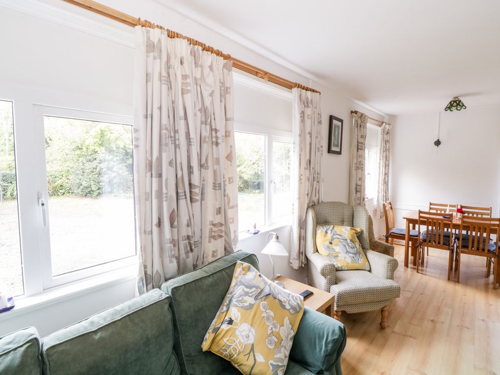 A living area with green sofa and patterned armchair near windows with curtains and a dining table with chairs in the background at Lakeside in Cahir near Ballinrobe County Mayo
