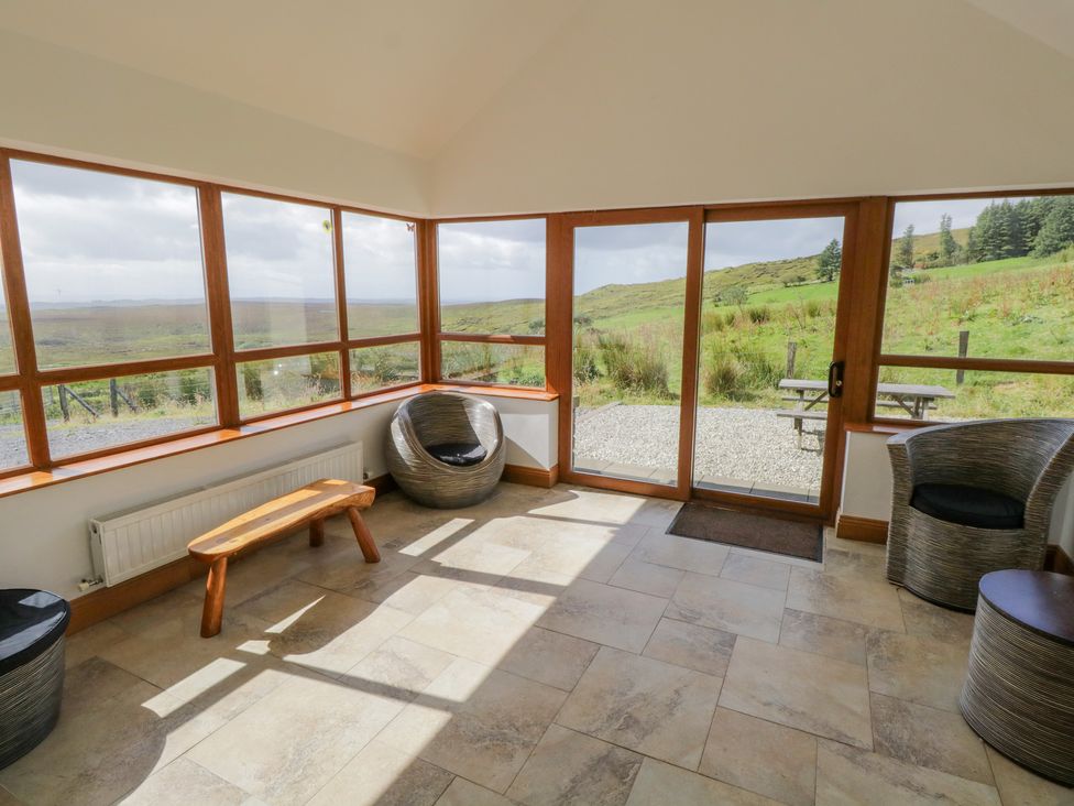 A sunroom with large windows and glass doors two wicker chairs a wooden bench and a view of green hills at Blue Stack House Meenaguish Beg near Donegal Town County Donegal