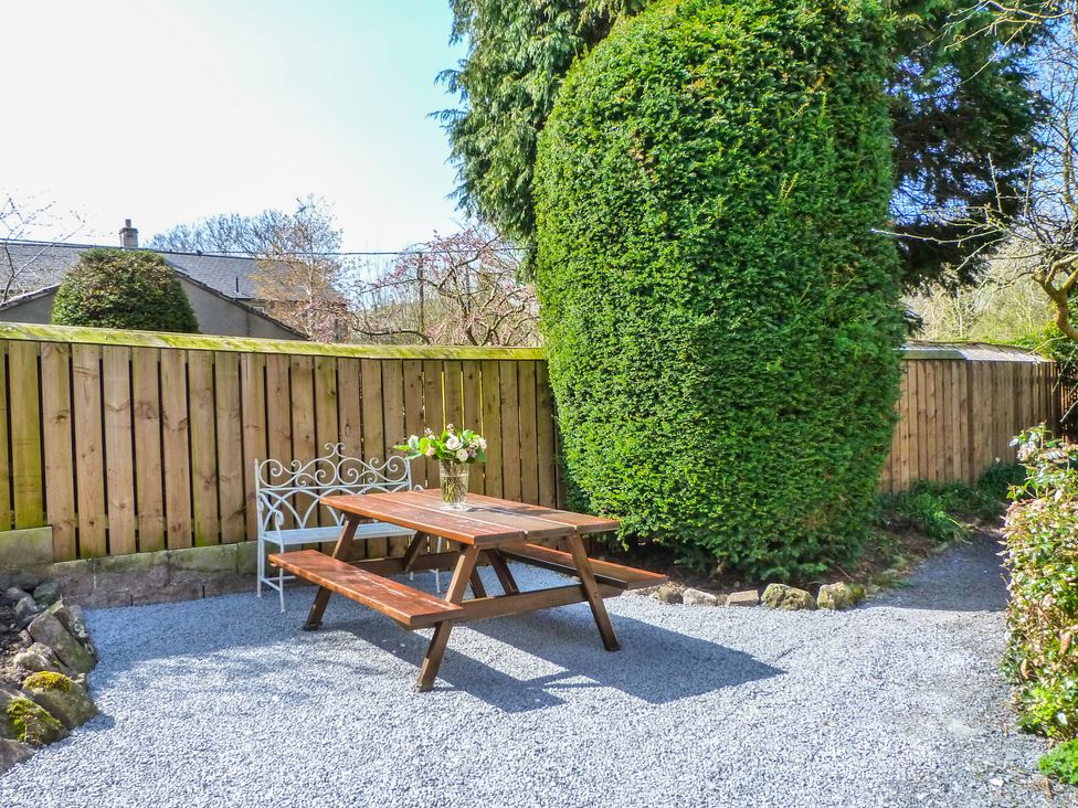 An outdoor seating area with a wooden picnic table and a white metal bench on gravel with a large trimmed hedge and wooden fence surrounding the area at Whitefriars Lodge in Settle