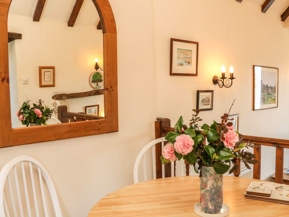 A dining area with a wooden table holding a vase of pink flowers a mirror reflecting a hallway and framed pictures on the wall at Primrose Cottage in Huntshaw near Great Torrington