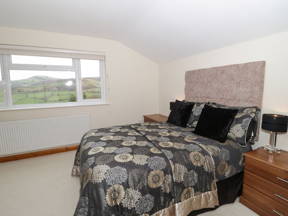 A bedroom with a double bed with patterned bedspread and bedside tables with a lamp and window showing a view of hills at Bryn Derw Holiday Cottage in Eglwysbach near Llanrwst and Conwy