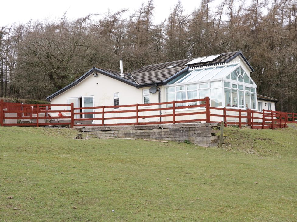 A single-story house with a glass conservatory and wooden fence on a grassy hill with trees in the background at Bryn Derw Holiday Cottage in Eglwysbach near Llanrwst and Conwy