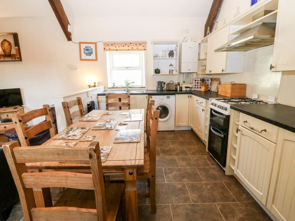 A kitchen and dining area with wooden chairs and table a stove and washing machine at Bwthyn y Dderwen Oak Cottage in Pwllheli