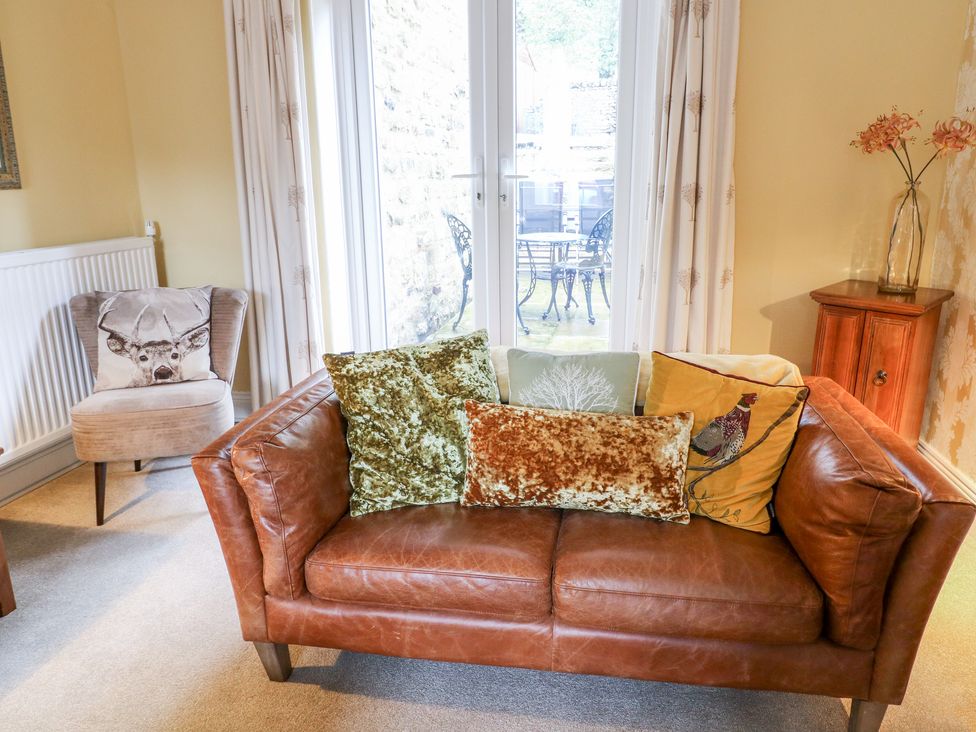 A brown leather sofa with five cushions in front of glass doors and a beige armchair with a deer cushion in a living room at Fern House in Kettlewell