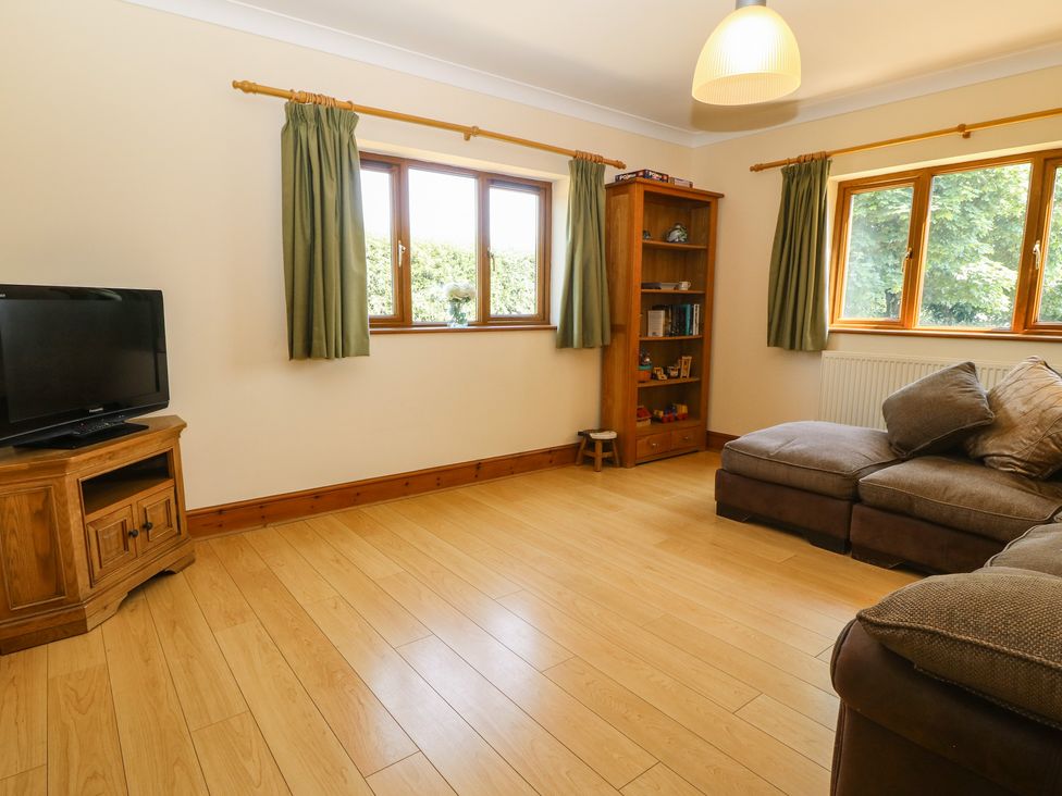 A living room with a television on a wooden stand a wooden bookshelf a grey sectional sofa and green curtains at Fferam Y Llyn in Rhosgoch near Cemaes Bay