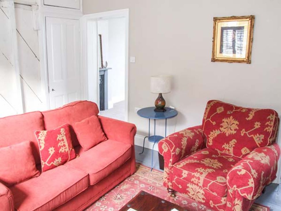 A living room with a red sofa and patterned armchair a round side table with a lamp and a framed picture on the wall at Woodland Cottage in Malvern