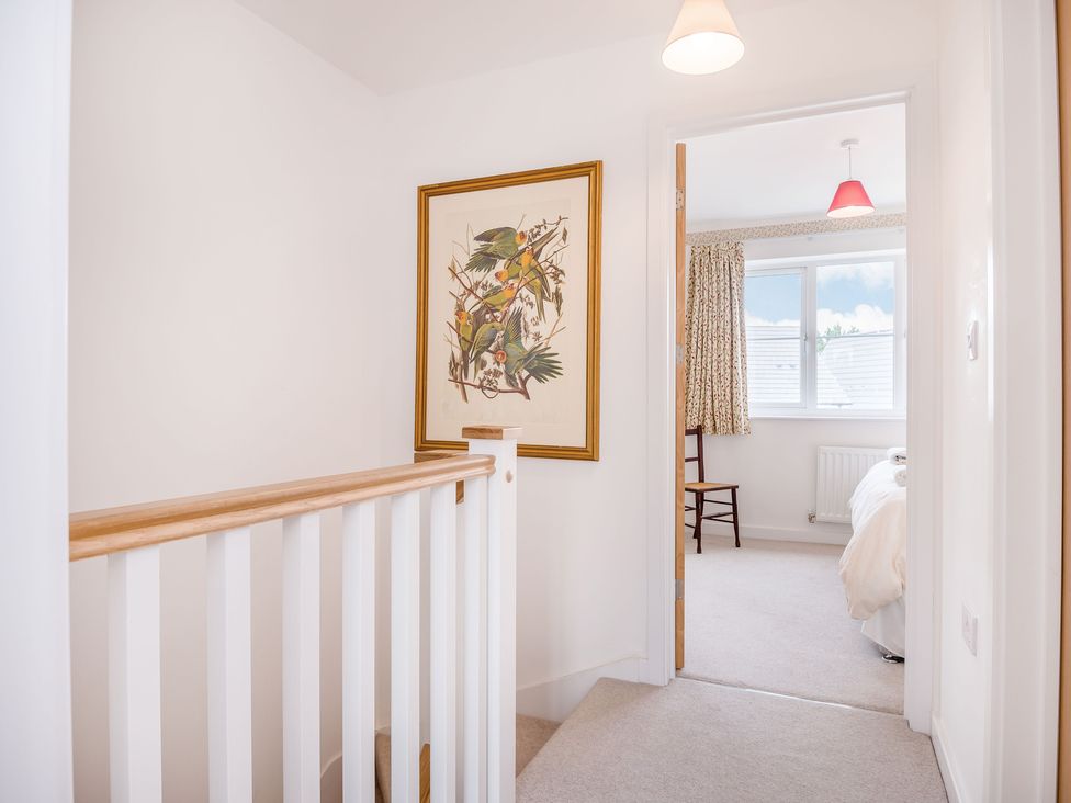 A hallway with a wooden railing and a framed bird print on the wall leading to a bedroom with a chair and a window at Saplings in Wells
