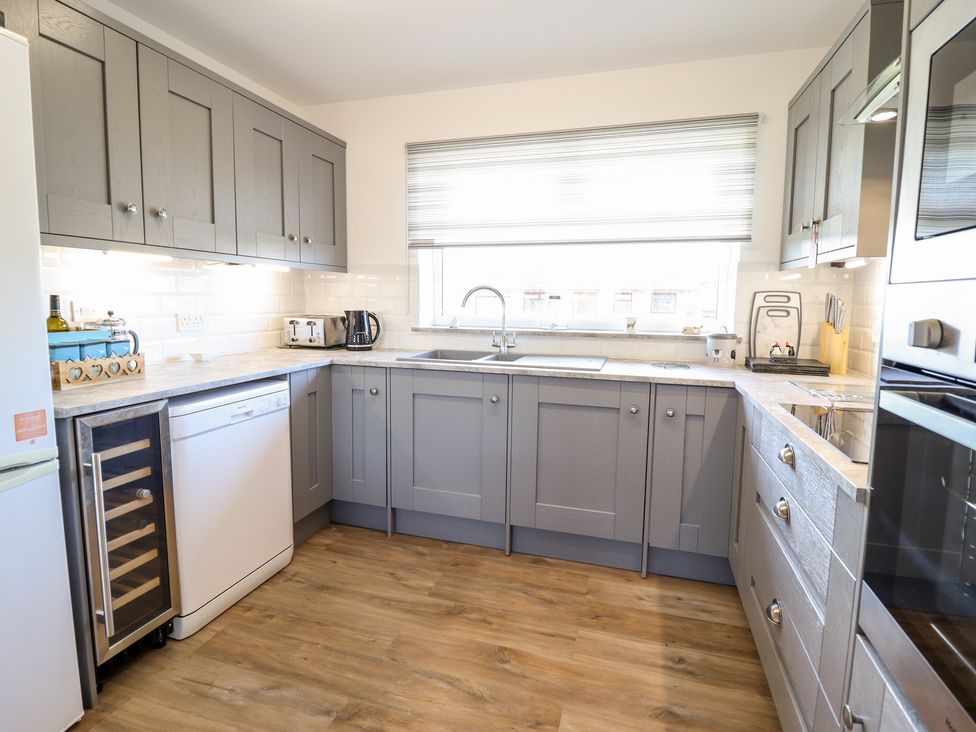 A kitchen with grey cabinets a dishwasher a wine cooler a toaster a kettle and a window with a blind at Sealyons Fairbourne near Dolgellau