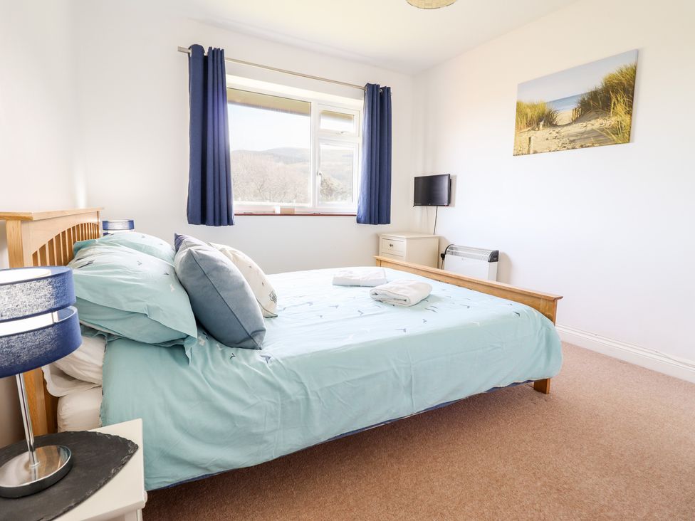 A bedroom with a wooden bed, blue bedding, pillows, a nightstand with lamp, a window with blue curtains, a small wall-mounted TV and a beach painting at Sealyons in Fairbourne near Dolgellau