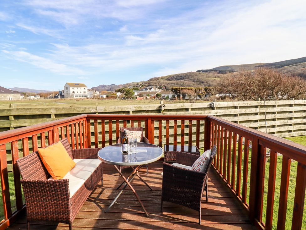 An outdoor wooden deck with wicker seating and a glass table with a bottle and two glasses at Sealyons Fairbourne near Dolgellau