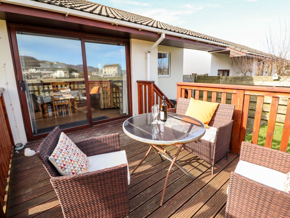 A wooden outdoor deck with a round glass table and wicker chairs with cushions at Sealyons Fairbourne near Dolgellau