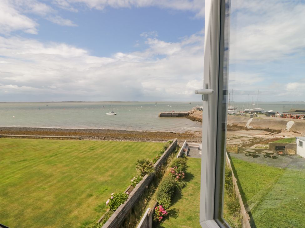 A view of a grassy yard with flowers and bushes by a beach and harbor visible through an open window at Driftwood 23 Roa Island in Roa Island